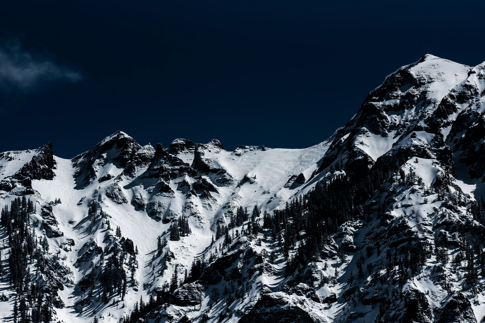 Jagged snow-covered mountain peaks with dark pine trees on the slopes against a deep blue sky